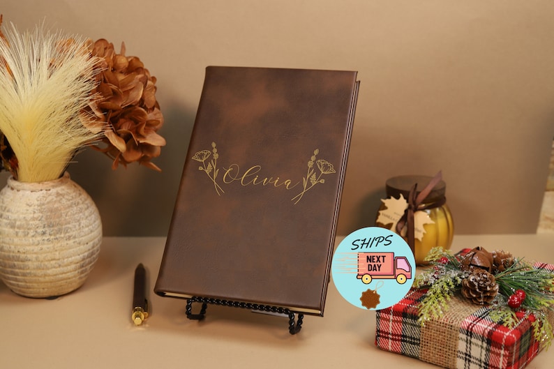 May include: A brown leather-bound journal with the name "Olivia" embossed in gold lettering. The journal is decorated with a gold floral design and is displayed on a black stand.
