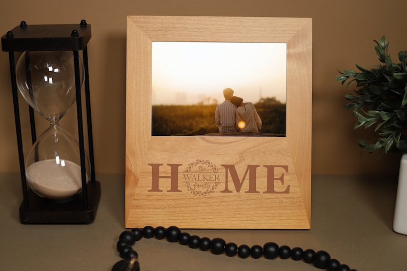 May include: A wooden picture frame with a photo of a couple sitting on a hill at sunset. The frame is engraved with the words "HOME" and "The Walker Family".