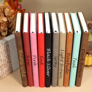 May include: A row of ten leather-bound journals in various colors, including rustic gold, rose, pink, red, black silver, gray, light brown, rawhide, teal, and dark brown. Each journal has a different color and is labeled with its color name.