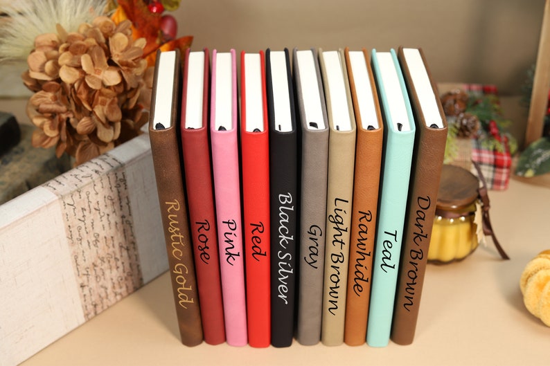 May include: A row of ten leather-bound journals in various colors, including rustic gold, rose, pink, red, black silver, gray, light brown, rawhide, teal, and dark brown. Each journal has a different color and is labeled with its color name.