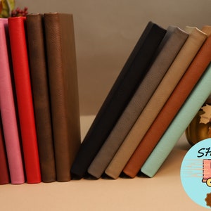 May include: A collection of ten leather-bound journals in various colors, including pink, red, brown, black, gray, beige, orange, and blue. The journals are arranged in a staggered pattern, with the blue journal at the front.