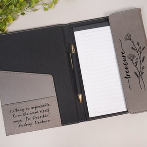 May include: A gray leather folder with a black pen and a notepad. The folder has a quote from Audrey Hepburn on the front: "Nothing is impossible. Even the word itself says: 'I'm Possible.' - Audrey Hepburn". The back of the folder has a floral design and the word "treasure" written in cursive.