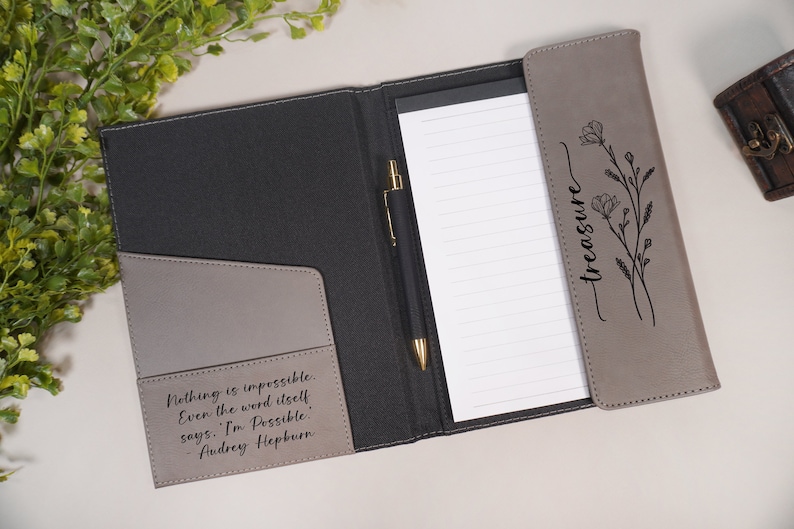 May include: A gray leather folder with a black pen and a notepad. The folder has a quote from Audrey Hepburn on the front: "Nothing is impossible. Even the word itself says: 'I'm Possible.' - Audrey Hepburn". The back of the folder has a floral design and the word "treasure" written in cursive.