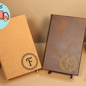 May include: Two personalized journals, one with a cork cover and the other with a brown leather cover. Both journals feature a gold-colored wreath with a letter in the center. The cork journal has the letter "F" and the leather journal has the letter "E" with the name "Ethan" written below it.