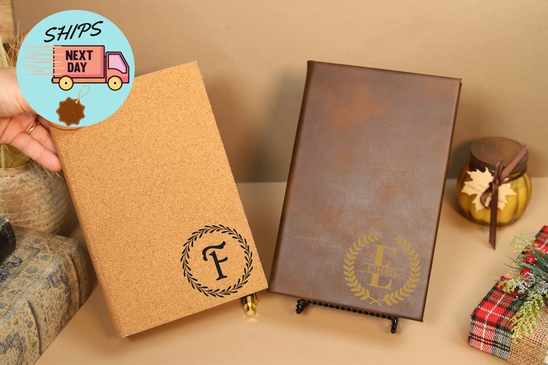 May include: Two personalized journals, one with a cork cover and the other with a brown leather cover. Both journals feature a gold-colored wreath with a letter in the center. The cork journal has the letter "F" and the leather journal has the letter "E" with the name "Ethan" written below it.