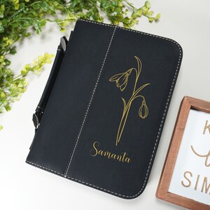 Op de afbeelding: Een zwart lederen dagboek met een gouden bloemenontwerp en de naam Samantha gegraveerd op de voorkant. Het dagboek heeft een pennenlus en een handvat.