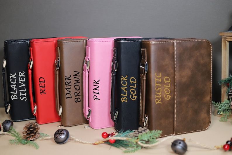 Op de afbeelding: Zeven verschillende gekleurde kunstleren bijbelhoezen met gouden letters. De kleuren zijn zwart, zilver, rood, donkerbruin, roze, zwart goud en rustiek goud.