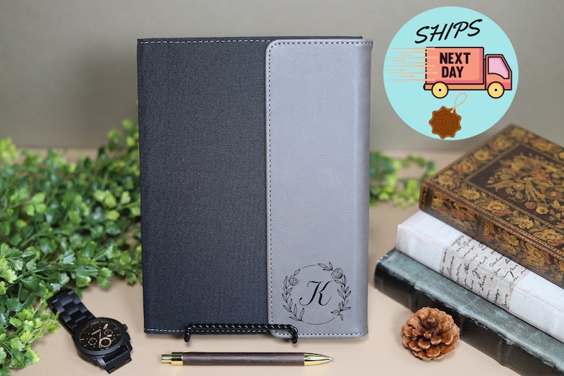 May include: A gray and black leather journal with a floral design and the letter "K" engraved on the front cover. The journal is closed and sitting on a table with a pen and a watch.