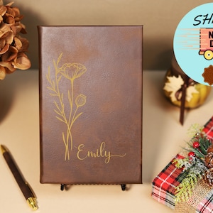 May include: A brown leather journal with a gold floral design and the name "Emily" engraved on the front. The journal is on a black stand.