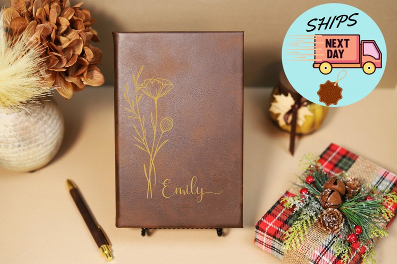 May include: A brown leather journal with a gold floral design and the name "Emily" engraved on the front. The journal is on a black stand.