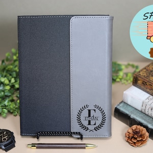 May include: A black and gray leather-bound journal with a personalized monogram of the letter "E" surrounded by a laurel wreath and the name "Ethan" engraved on the front cover.