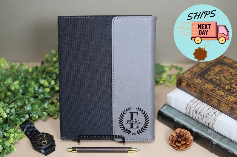 May include: A black and gray leather-bound journal with a personalized monogram of the letter "E" surrounded by a laurel wreath and the name "Ethan" engraved on the front cover.