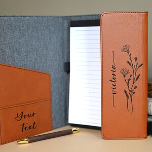 May include: A brown leather folder with a gray fabric exterior. The folder has a pocket on the front with the text "Your Text" engraved on it. The inside of the folder has a notepad with the text "victoria" engraved on it, along with a floral design.