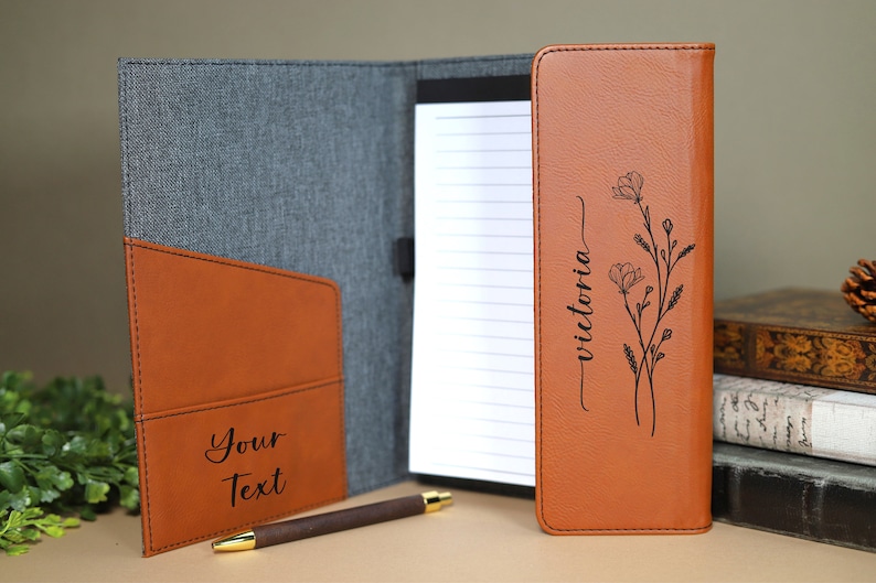 May include: A brown leather folder with a gray fabric exterior. The folder has a pocket on the front with the text "Your Text" engraved on it. The inside of the folder has a notepad with the text "victoria" engraved on it, along with a floral design.