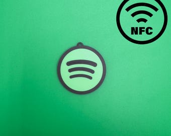Pode incluir: Um pingente verde com o logotipo do Spotify e um símbolo NFC preto sobre um fundo verde. O logotipo do Spotify é um círculo verde com três linhas curvas pretas. O símbolo NFC tem o texto "NFC" abaixo.