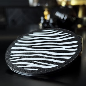 May include: A circular coaster featuring a high-contrast black and white zebra stripe pattern. The surface has a textured finish, and the design consists of horizontal, wavy white lines set against a solid black background across the round disc.
