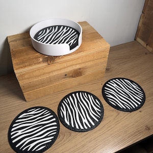 May include: A set of three round coasters featuring a black and white zebra stripe design. A white coaster holder with the same pattern is also included. The coasters and holder are on a wooden surface.