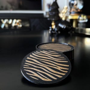 May include: A set of two circular coasters featuring a black base with a textured gold zebra stripe pattern. The coasters are stacked, with one positioned flat in the foreground and the other partially visible behind it, showcasing a consistent design.