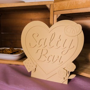 Può includere: Un cartello a forma di cuore beige con le parole "Salty Bar" ritagliate. Il cartello presenta dettagli di pretzel, fragole e rose. Il cartello è su una superficie viola, con casse di legno sullo sfondo.