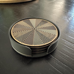 May include: A stack of circular coasters with a geometric starburst pattern in black and natural wood tones, housed within a matte black circular holder. The top coaster features intricate, laser-cut concentric lines radiating from the centre.