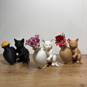 Könnte beinhalten: Eine Sammlung von katzenförmigen Vasen in Schwarz, Weiß und Beige, jede mit bunten Blumen. Die Vasen sind auf einer Holzoberfläche angeordnet und bilden eine dekorative Präsentation. Die Blumen umfassen gelbe, rosa und rote Blüten.