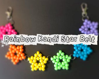 以下が含まれることがあります： ピンク、オレンジ、黄色、緑、青、紫の星型のビーズが付いたレインボーキャンディスターベルト。「Rainbow Kandi Star Belt」の文字が白い背景に表示されています。