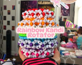 以下が含まれることがあります： カラフルなレインボーキャンディローテーターのブレスレットが手に持たれています。ブレスレットは、赤、オレンジ、白、青、紫、ピンクなどの色のビーズで作られています。「Rainbow Kandi Rotator」という文字が画像に表示されています。