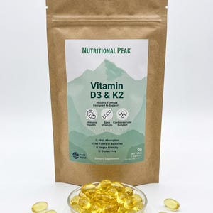 May include: A brown kraft paper pouch of Nutritional Peak Vitamin D3 & K2 dietary supplement with a mountain graphic label. The label lists 90 softgels and benefits for immune, bone, and cardiovascular health. A clear dish of yellow softgels sits in front.