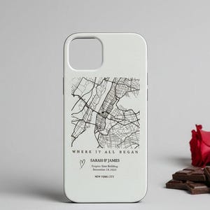 May include: White phone case featuring a black and white map of New York City. The case includes the text "WHERE IT ALL BEGAN", a heart symbol, names, and a date. Red roses and chocolate bars are in the background.