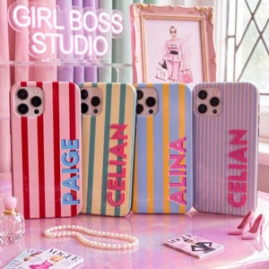 May include: Four colorful phone cases with vertical stripes and names in bold letters. The cases are red, yellow, blue, and pink. The image also includes a neon sign that says "GIRL BOSS STUDIO".