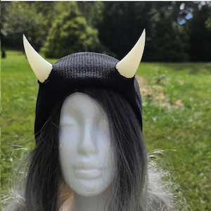 May include: Black beanie with two white horns attached to the top. The beanie is made of a soft, knitted fabric.