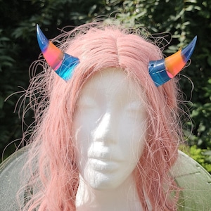 May include: A pair of colorful, iridescent devil horns in blue, orange, and pink hues, attached to a pink wig on a mannequin head. The horns are curved and have a textured appearance, perfect for cosplay or costume events.