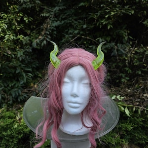 May include: A pink wig with long, wavy hair is styled on a white mannequin head. Two translucent, neon green horns are attached to the wig. The mannequin is mounted on a gray stand, with a green, circular collar.