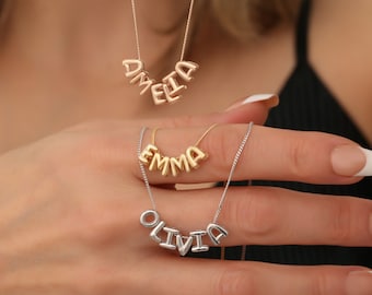 May include: Three gold necklaces with personalised names, Amelia, Emma, and Olivia, spelled out in individual letters. The necklaces are on a model's hand.