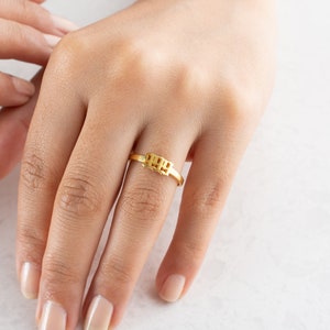 May include: A gold-colored ring with the numbers "1999" in a block design. The ring is on a finger, with the hand and other fingers visible. The ring is a simple band with the numbers in a rectangular shape.