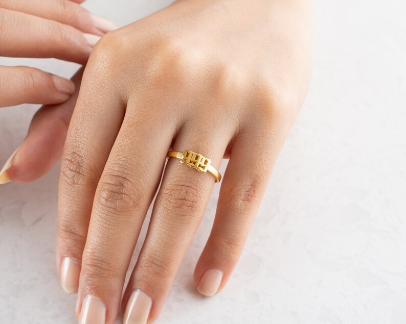 May include: A gold-colored ring with the numbers "1999" in a block design. The ring is on a finger, with the hand and other fingers visible. The ring is a simple band with the numbers in a rectangular shape.