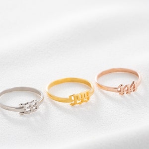 May include: Three delicate rings in silver, gold, and rose gold. Each ring features a numerical design: 333, 999, and 444. The rings are displayed on a white surface, showcasing their minimalist style and metallic shine.