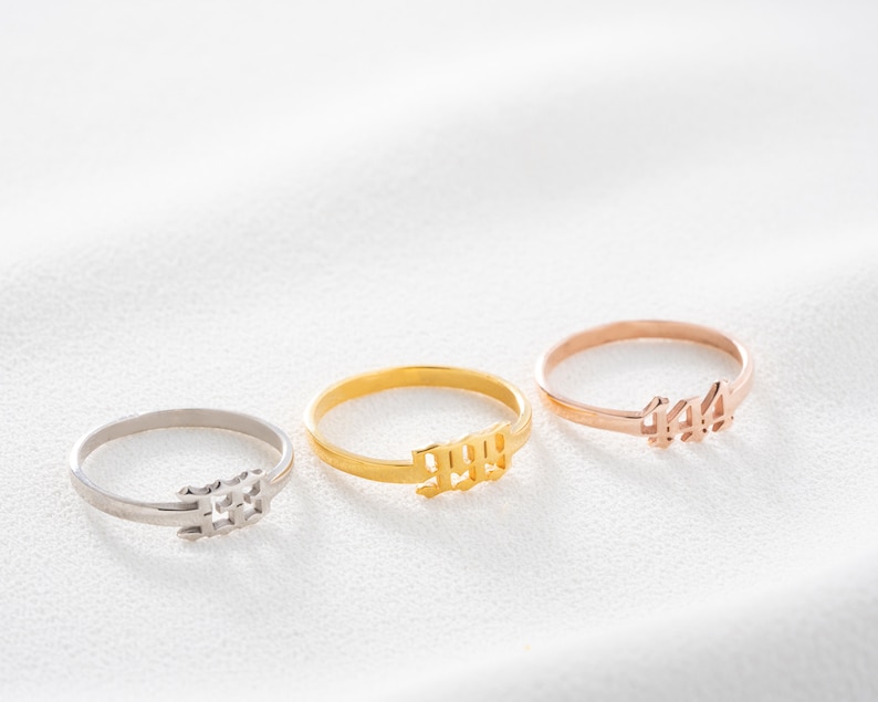 May include: Three delicate rings in silver, gold, and rose gold. Each ring features a numerical design: 333, 999, and 444. The rings are displayed on a white surface, showcasing their minimalist style and metallic shine.