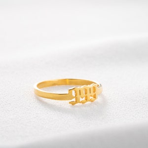 May include: A gold-colored ring with the number 1999 in a stylized font. The ring is set against a white background, highlighting its polished finish and design. The ring is a simple band with the numbers as a central feature.