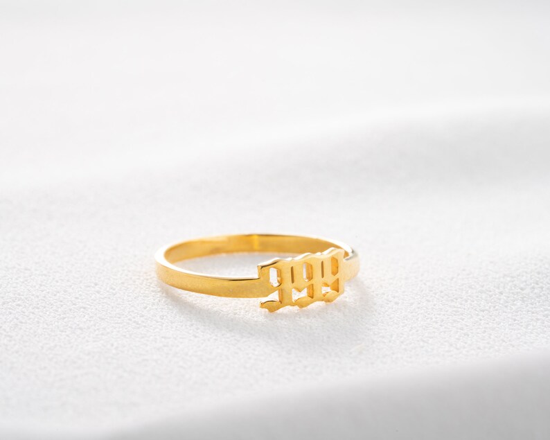 May include: A gold-colored ring with the number 1999 in a stylized font. The ring is set against a white background, highlighting its polished finish and design. The ring is a simple band with the numbers as a central feature.