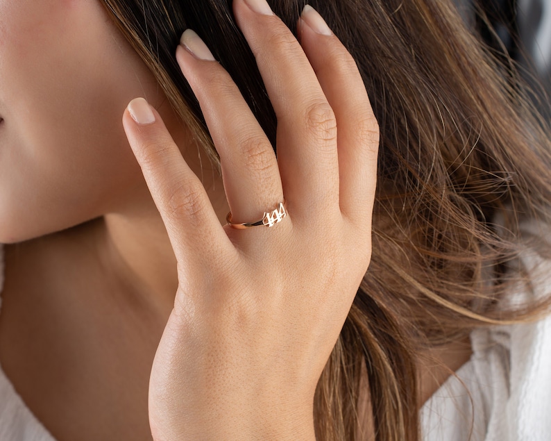 May include: A delicate gold ring featuring the number "444" in a minimalist design. The ring is worn on a finger, showcasing its slender band and the subtle, yet meaningful, numerical detail. The ring's warm gold tone contrasts with the skin.