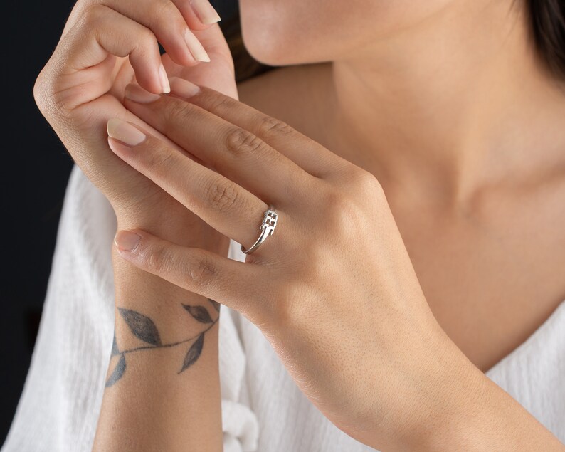 May include: A silver ring with a geometric design. The ring features a series of small, square-shaped cutouts arranged in a linear pattern. The ring is worn on a finger, showcasing its minimalist and modern aesthetic. The ring is a piece of jewelry.