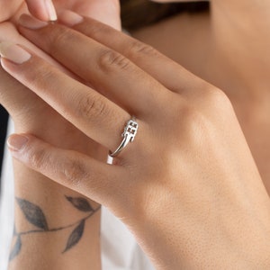 May include: A silver ring with a geometric design. The ring features a slender band that curves up to a square-shaped centerpiece with a cross-like pattern. The ring is worn on a finger, showcasing its delicate and modern aesthetic.