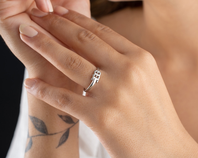 May include: A silver ring with a geometric design. The ring features a slender band that curves up to a square-shaped centerpiece with a cross-like pattern. The ring is worn on a finger, showcasing its delicate and modern aesthetic.
