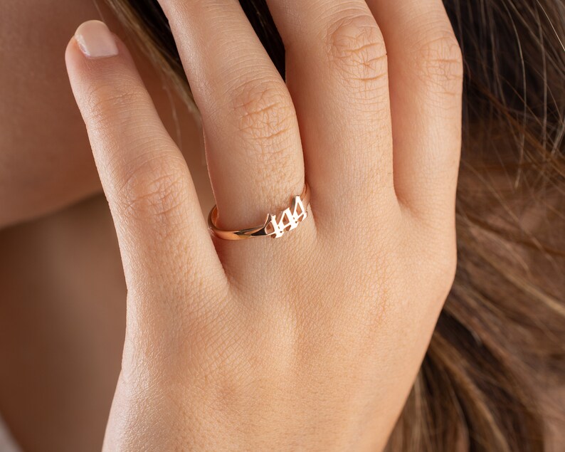 May include: A rose gold ring with the number 444 cut out in a delicate font. The ring is shown on a person's finger, highlighting its simple yet elegant design. The ring is a piece of jewelry.