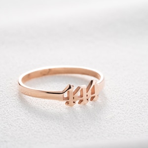 May include: Rose gold ring featuring the number 444 in a delicate, stylized font. The ring is thin and minimalist, with a polished finish. The ring is set against a white background.