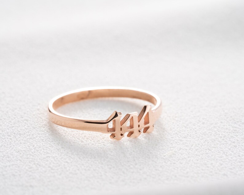 May include: Rose gold ring featuring the number 444 in a delicate, stylized font. The ring is thin and minimalist, with a polished finish. The ring is set against a white background.