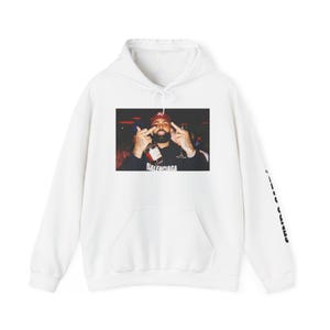 May include: White hooded sweatshirt with a front pocket and a graphic print. The graphic print features a person holding a bottle and making a hand gesture. The word "BALENCIAGA" is printed on the graphic. The sleeve has a black print.