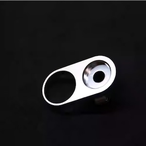 Könnte beinhalten: Eine silberfarbene Objektivabdeckung mit zwei kreisförmigen Öffnungen. Eine Öffnung ist ein größerer Kreis, die andere ein kleinerer Kreis. Die Abdeckung befindet sich auf schwarzem Hintergrund.