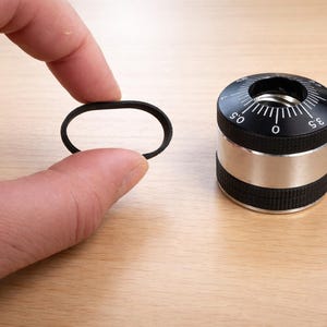 Peut inclure: Un objet cylindrique noir et argenté avec un cadran et des mesures, à côté d'un anneau ovale noir. L'objet a une bande argentée et des sections texturées noires. Le cadran a des marquages et des chiffres blancs.